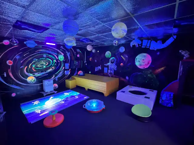 The Space room at The Special Lioness under UV light: a galaxy mural, hanging planets, a glowing floor projection and soft seating